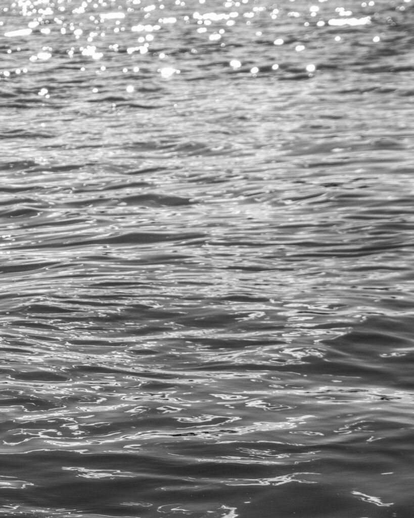 Black-and-white photograph of sunlight sparkling across rippling water, with bright reflections fading into darker, more defined ripples.