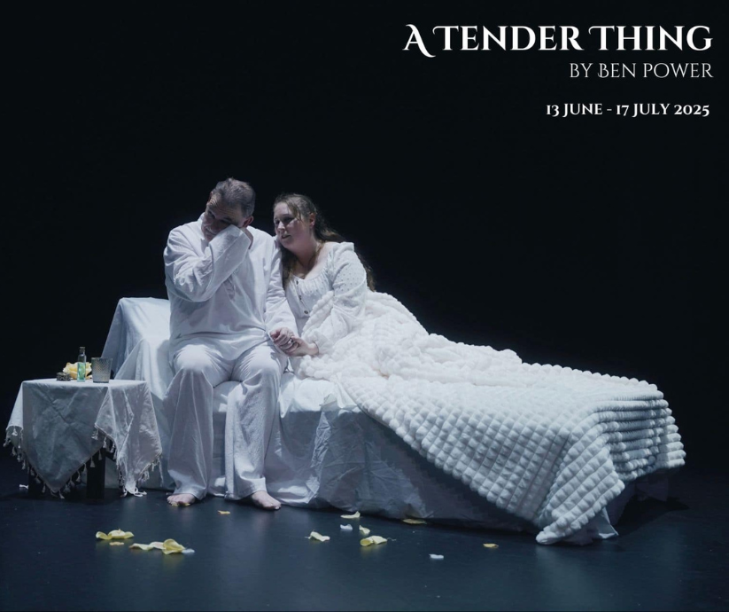 Two performers dressed in white sit together on the edge of a bed under cool stage lighting in A Tender Thing (2025).