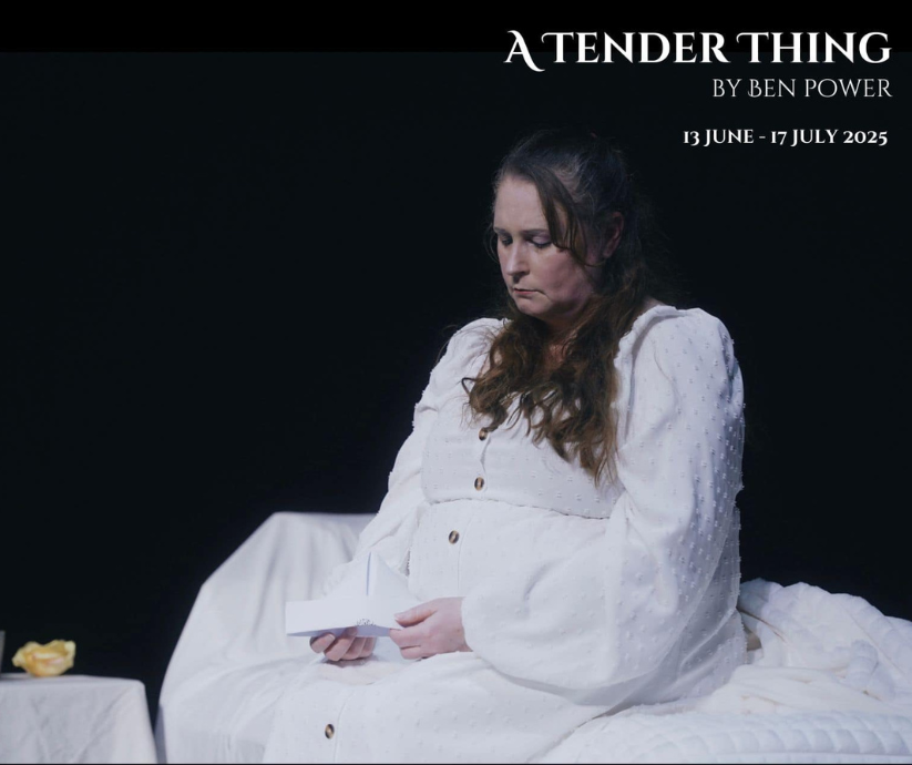 Close-up of a performer in a white dress reading a paper while seated on a bed under soft stage lighting in A Tender Thing (2025).