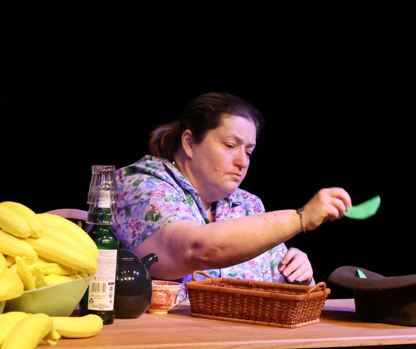 Amanda Sutherland as Kath in The Banana Bender, examining a green prop at a table surrounded by yellow banana props.