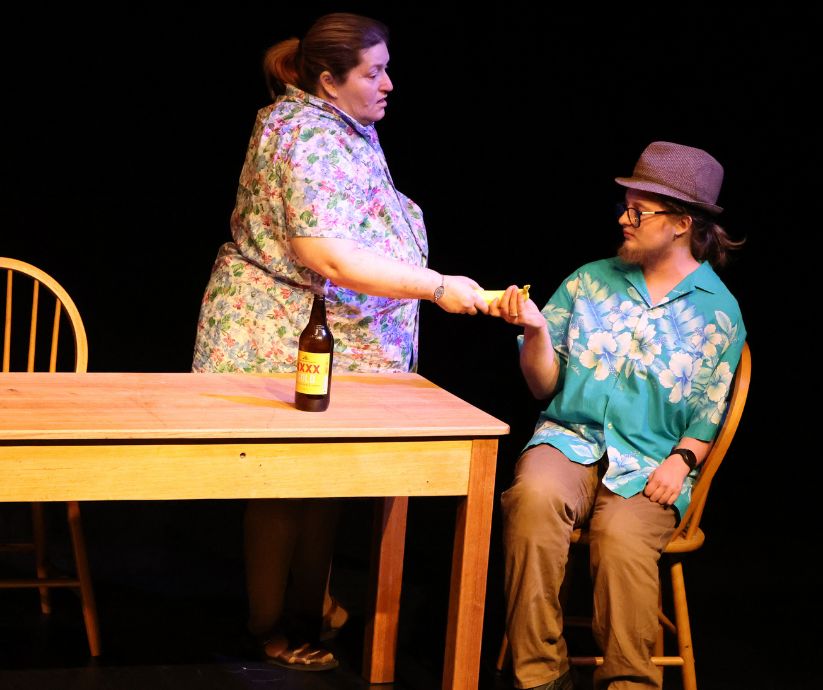 Amanda Sutherland as Kath standing beside Elliot Howe as Len, offering him a small prop during a scene from The Banana Bender.