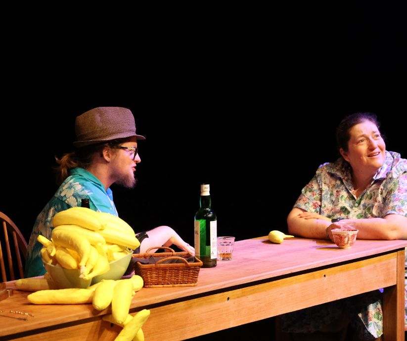 Elliot Howe as Len and Amanda Sutherland as Kath seated at a wooden table surrounded by yellow banana props during The Banana Bender.