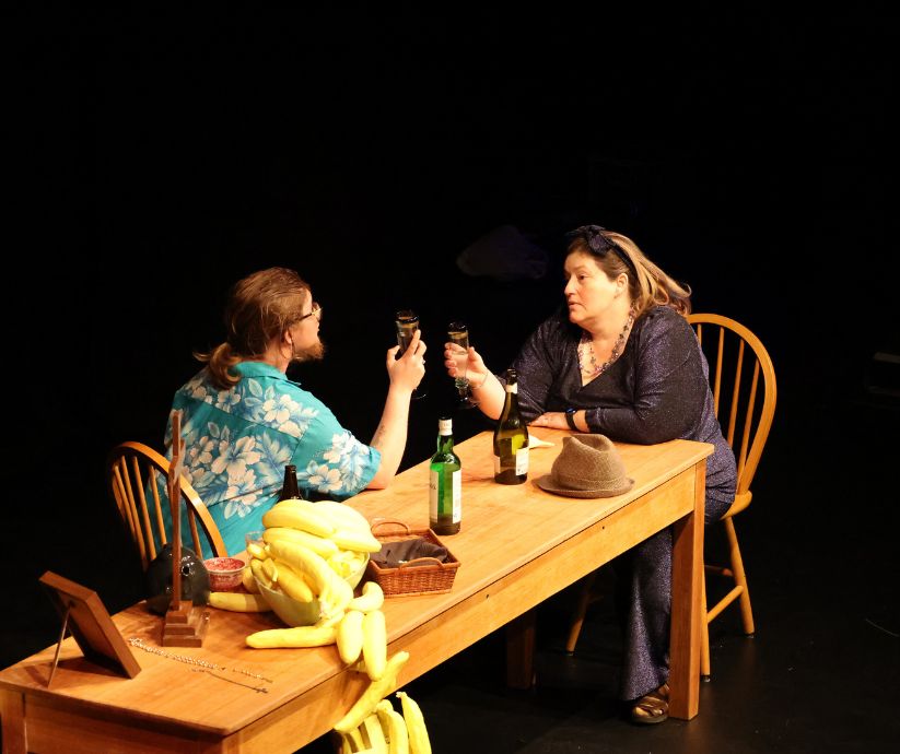Amanda Sutherland as Ruth and Elliot Howe as Len raising drinking glasses across a wooden table during The Banana Bender.