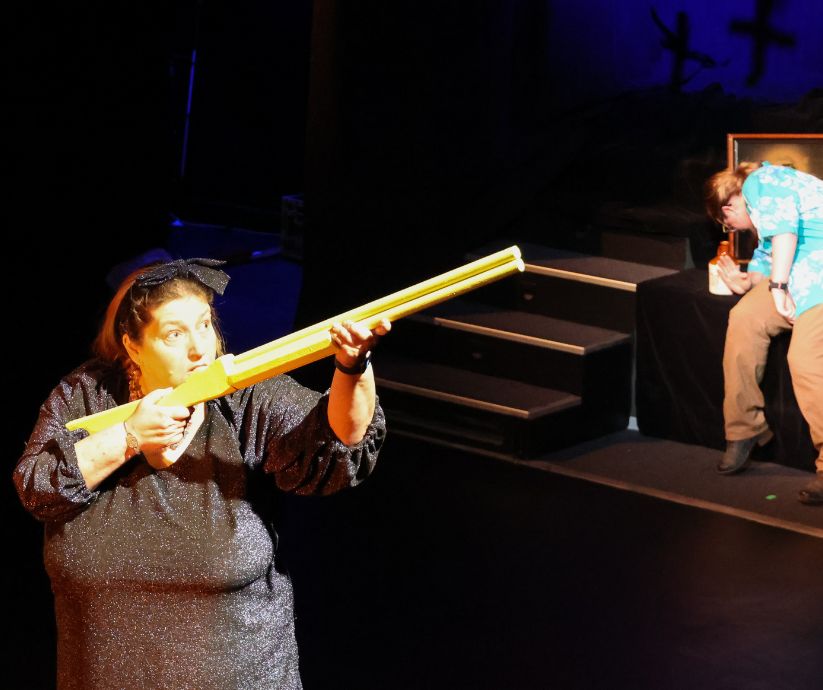 Amanda Sutherland as Ruth aiming a long yellow theatrical prop while Elliot Howe as Len reacts in the background during The Banana Bender.