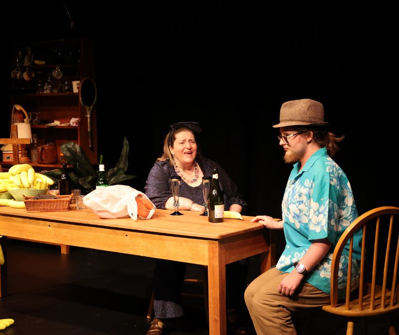Amanda Sutherland as Ruth talking with Elliot Howe as Len across a wooden table, with banana props and bottles visible on stage in The Banana Bender.