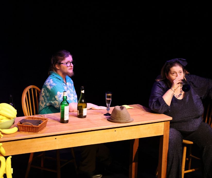Amanda Sutherland as Ruth drinking from a glass while Elliot Howe as Len sits beside her at a table during The Banana Bender.