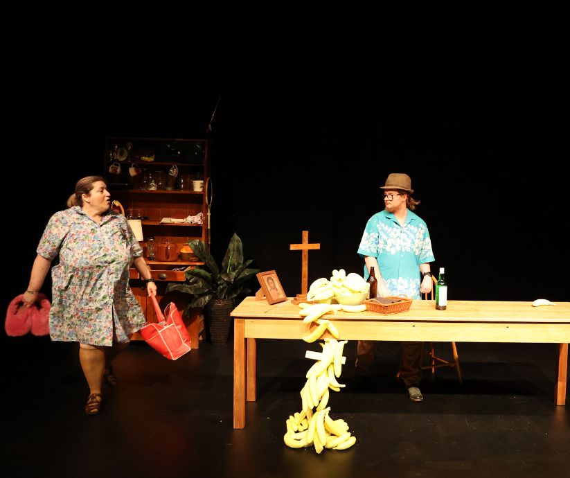 Amanda Sutherland as Kath walking across the stage carrying red bags while Elliot Howe as Len stands beside a table covered with banana props.