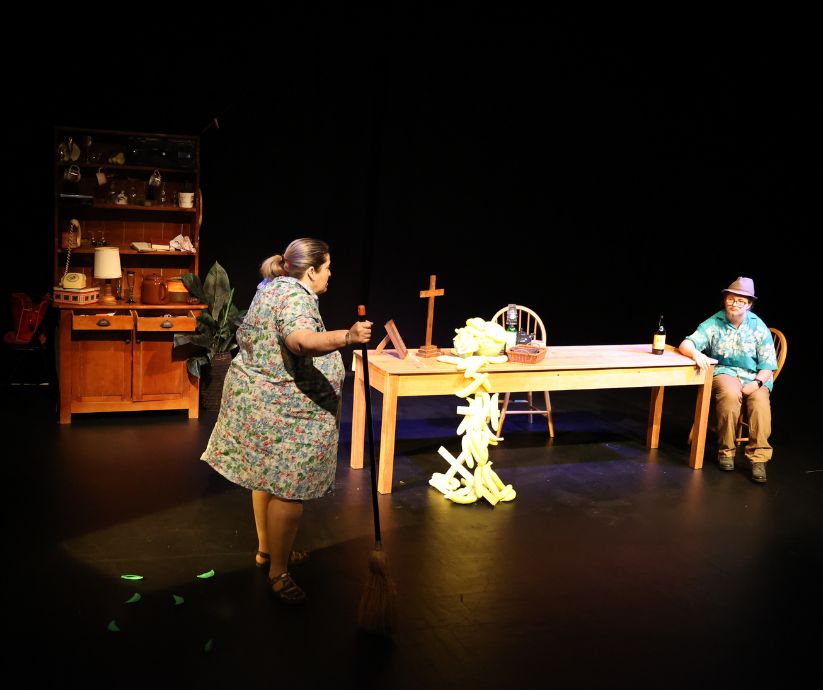 Amanda Sutherland as Kath holding a broom while facing Elliot Howe as Len across a wooden table in The Banana Bender.